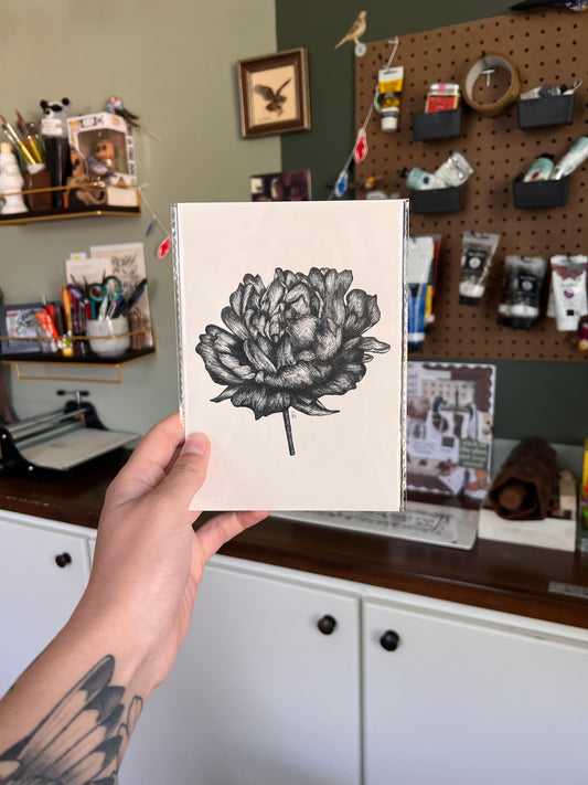 Hand holding a black and white floral illustration in a casual indoor setting.