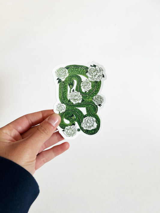 Hand holding a decorative green and white sticker with snake and floral patterns on a plain background