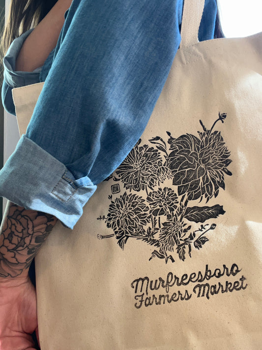 Beige tote bag with floral design and text, held by a person wearing blue jean shirt.