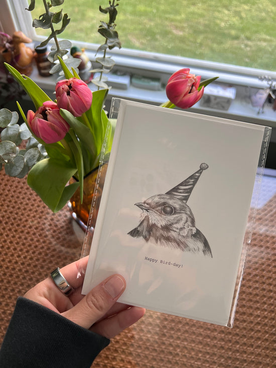 Card with a bird with a birthday hat illustration held by a hand next to pink tulips.