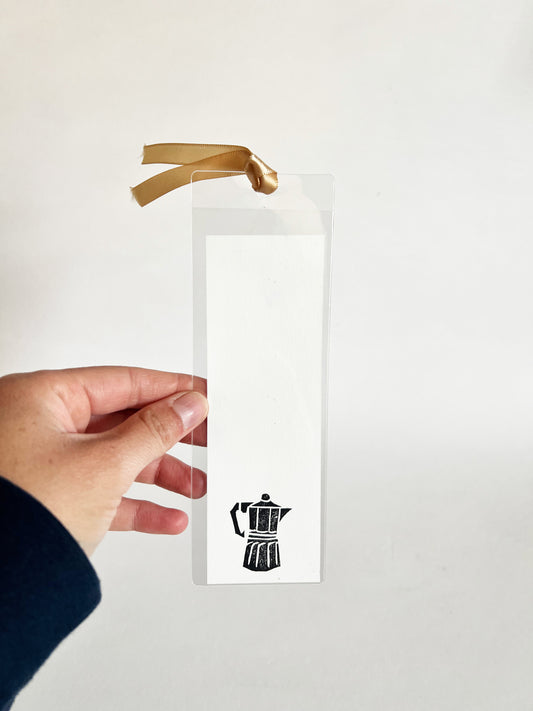 Hand holding a bookmark with a black coffee pot illustration on a white background