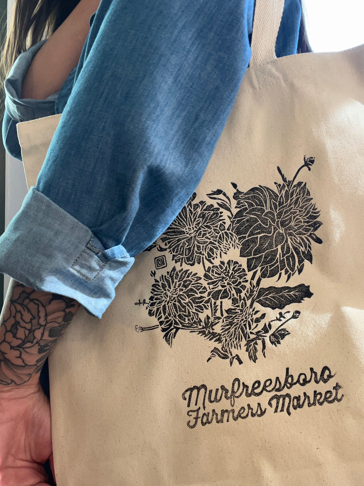 Beige tote bag with floral design and text, held by a person wearing blue jean shirt.