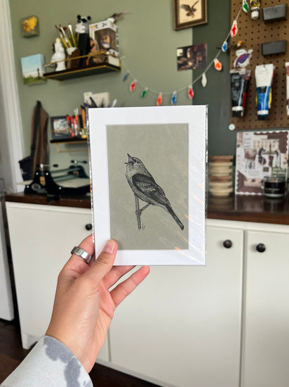 A hand holding a matted bird drawing.
