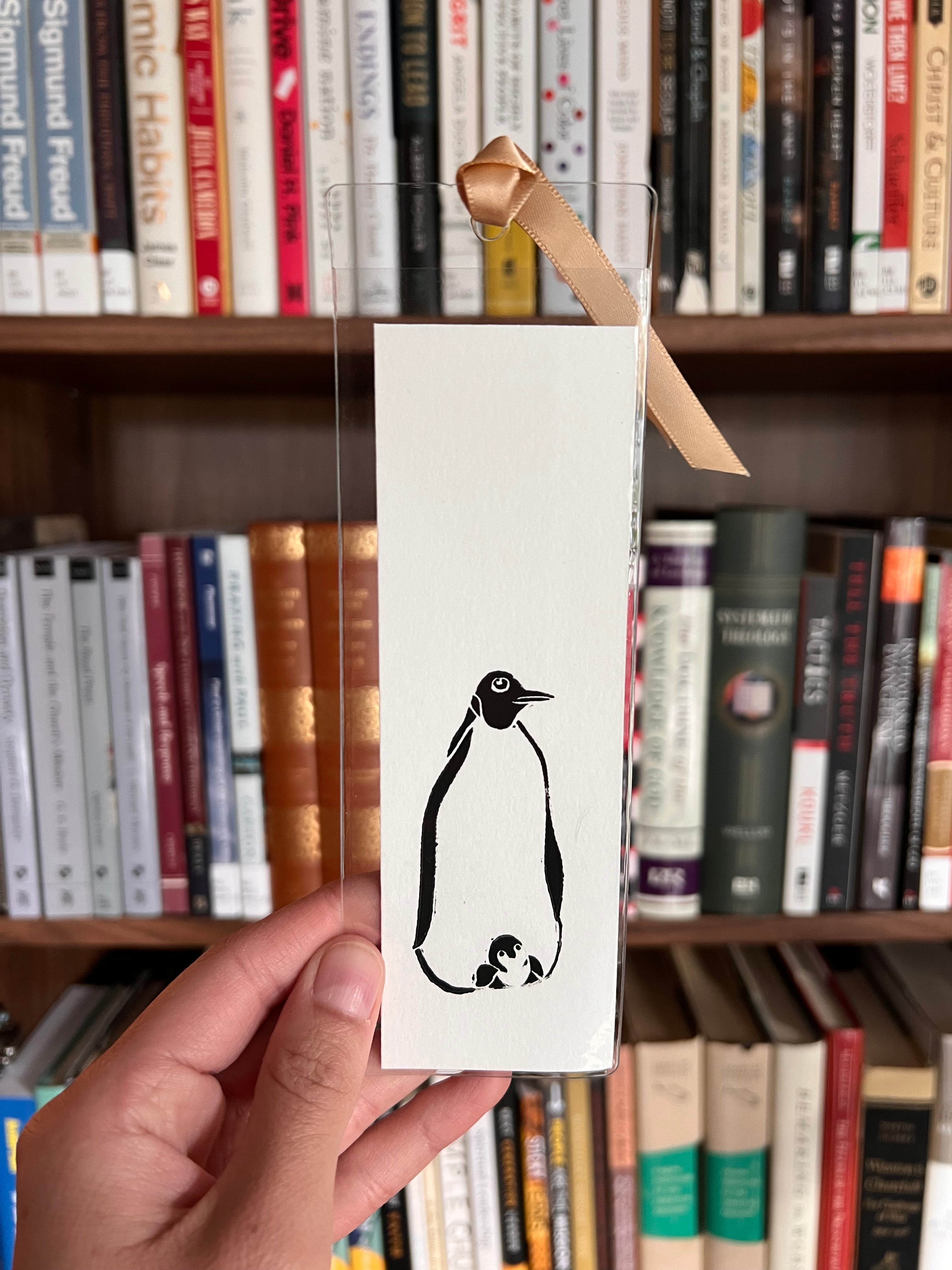 Hand holding a card with a black and white illustration of a penguin in front of a bookshelf.