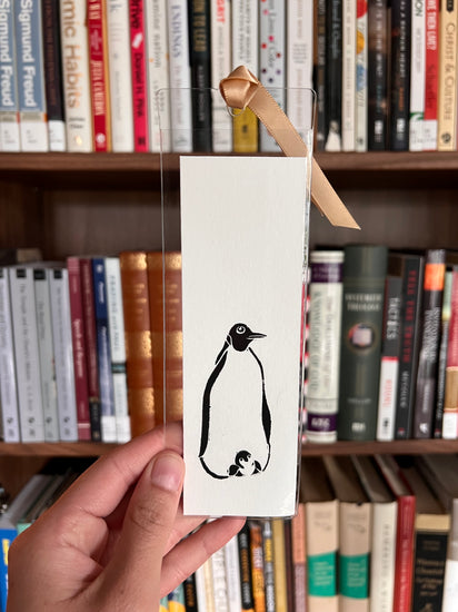 Hand holding a card with a black and white illustration of a penguin in front of a bookshelf.